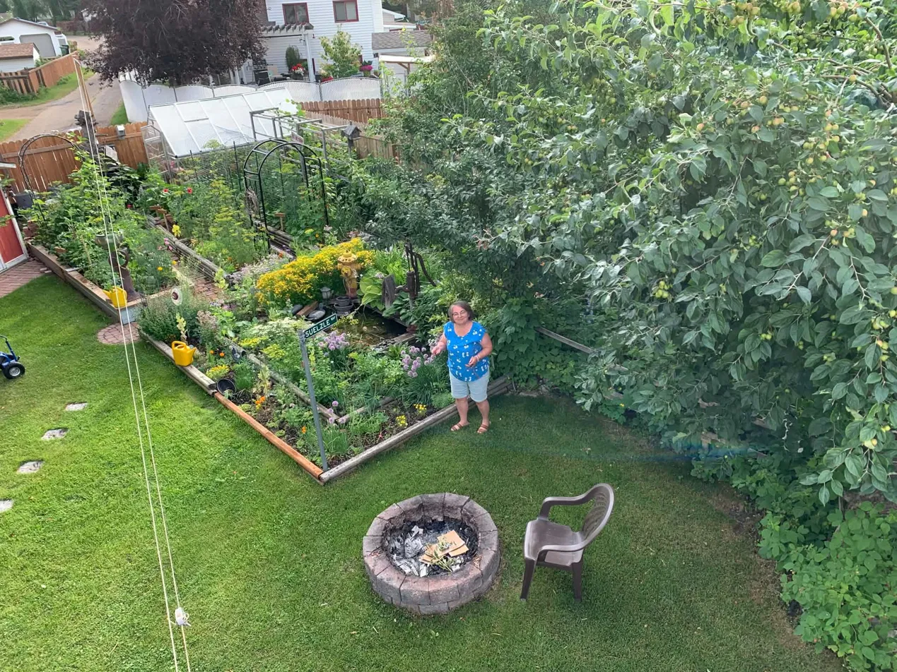 Cindy standing in her Edmonton backyard garden, with the Suelzle Way sign, greenhouse, apple trees, and raised garden beds visible.