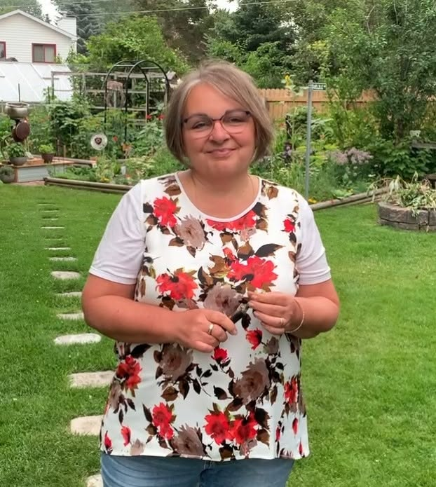 Cindy standing in her backyard garden with raised beds, stepping stones, and a greenhouse behind her